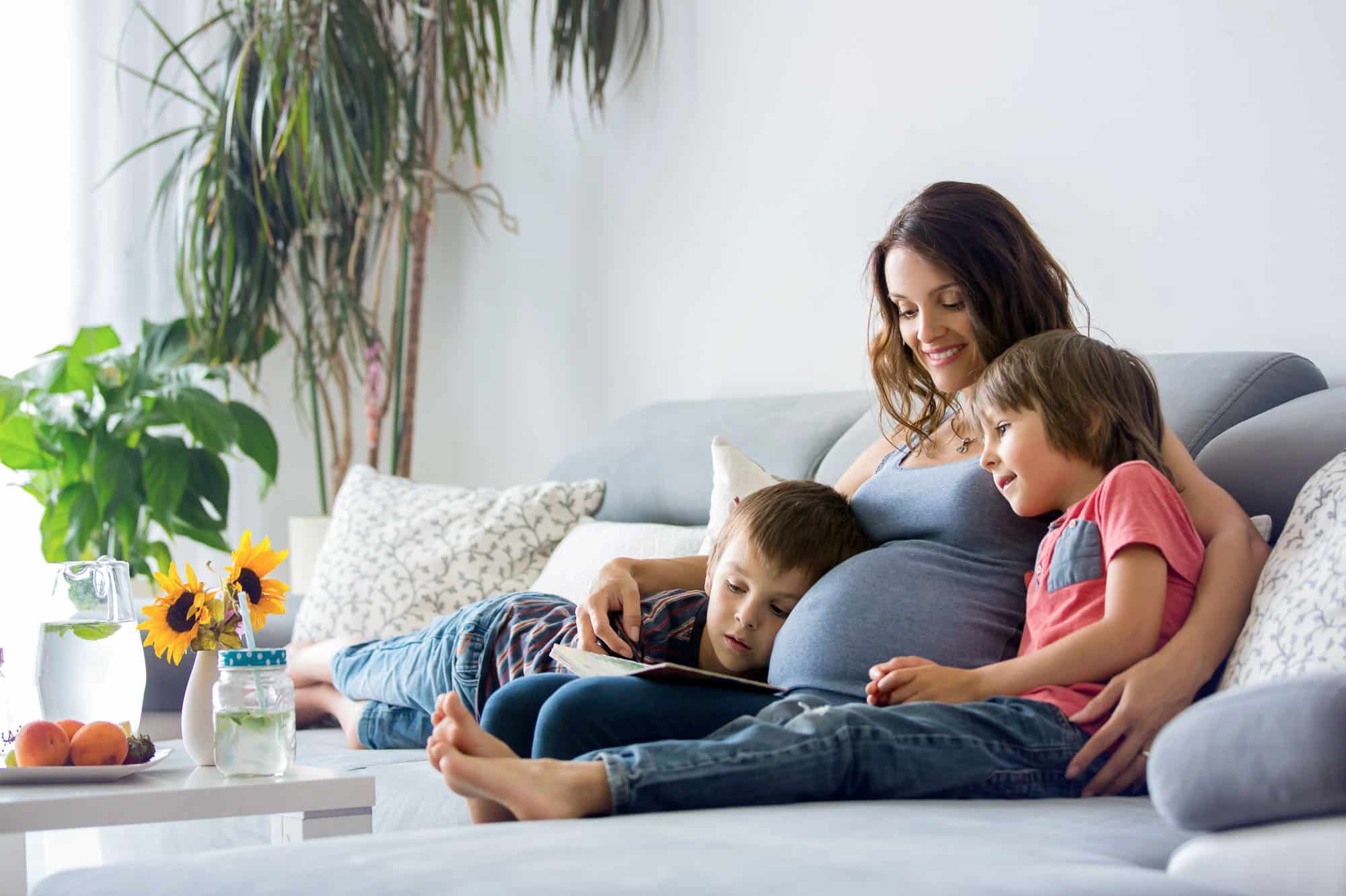 a pregnant mother sitting on a sofa and reading to her two young sons