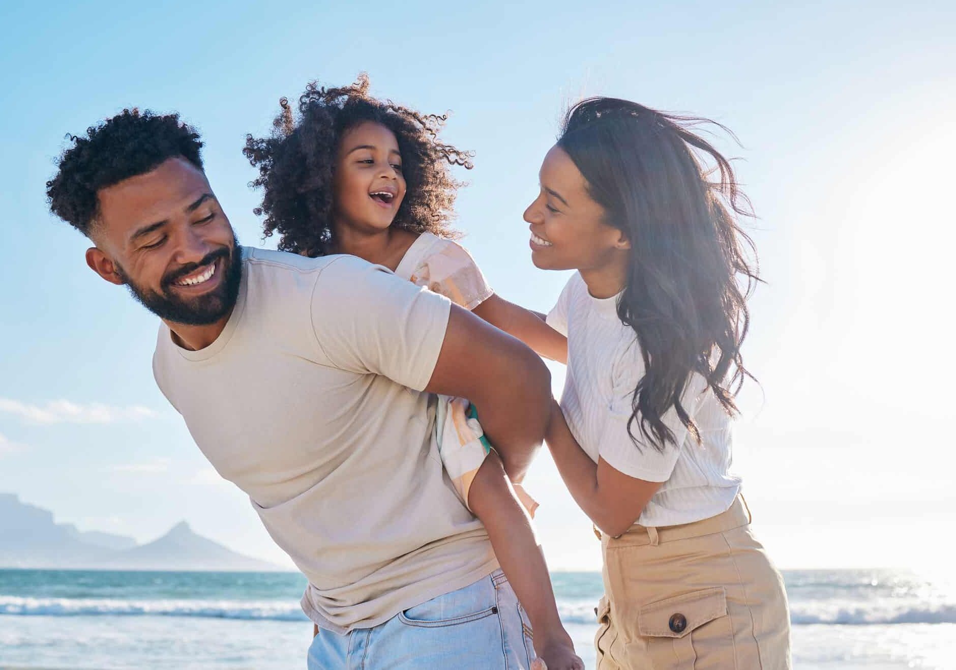 a dad giving his daughter a piggy back ride at the beach with the mom standing next to them