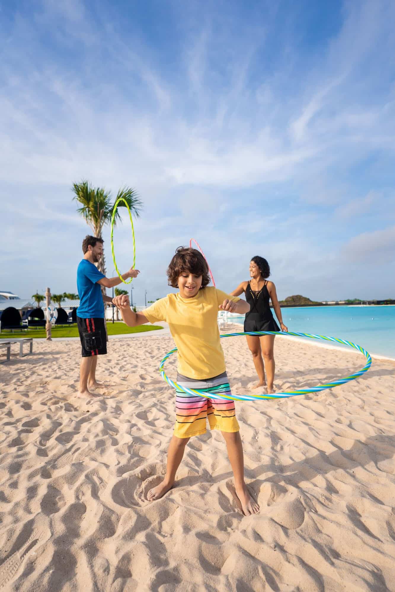 a boy hula hooping in the sand on the beach with a man and woman behind him
